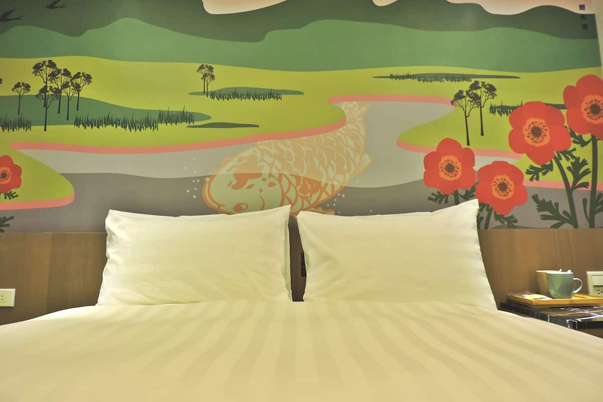 A hotel room with a bed, pillows, a nightstand with a cup and a book, and a wall mural depicting a landscape with flowers and birds.