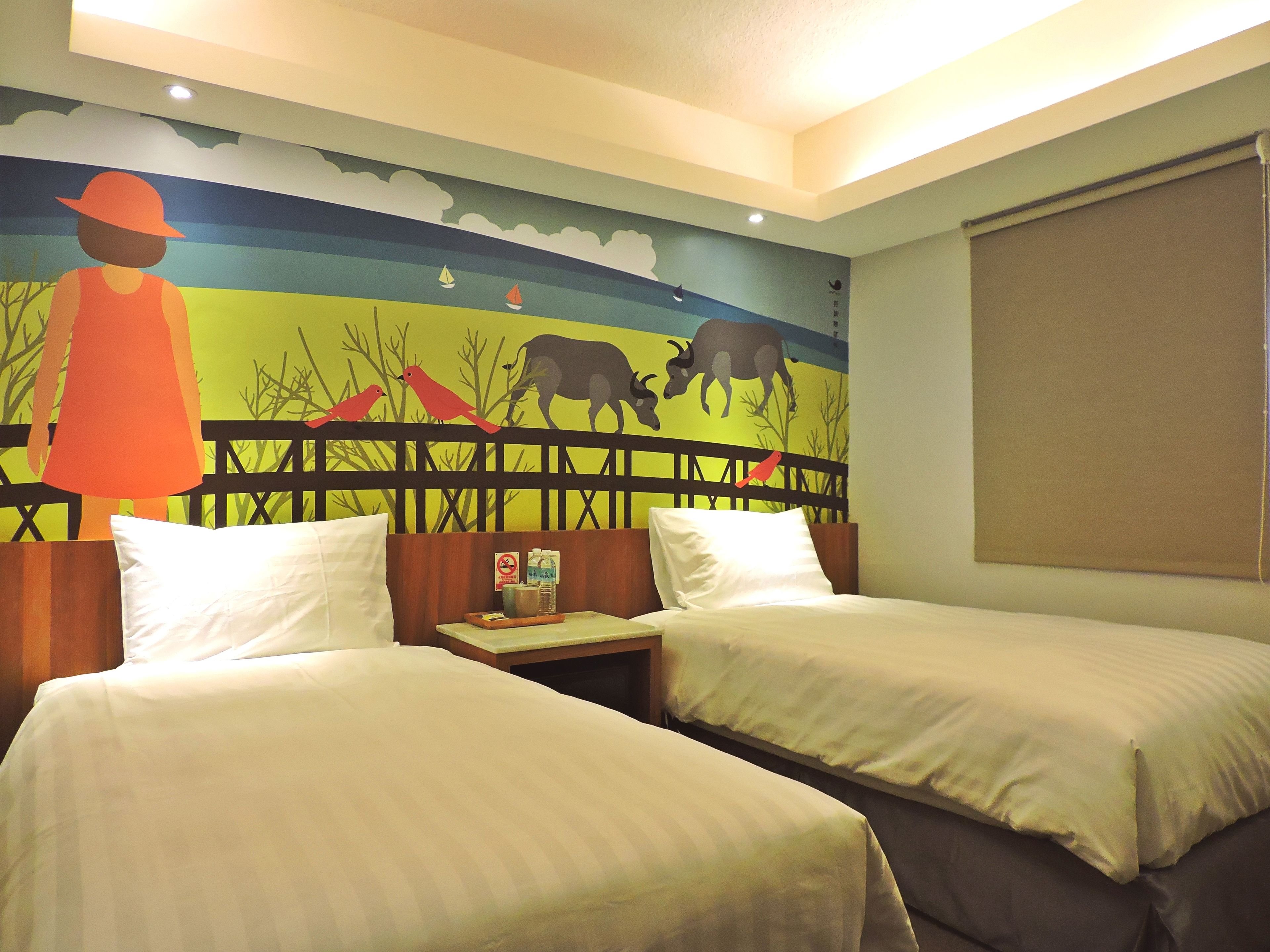 A hotel room with two beds, a mural on the wall, and a window with a blind.