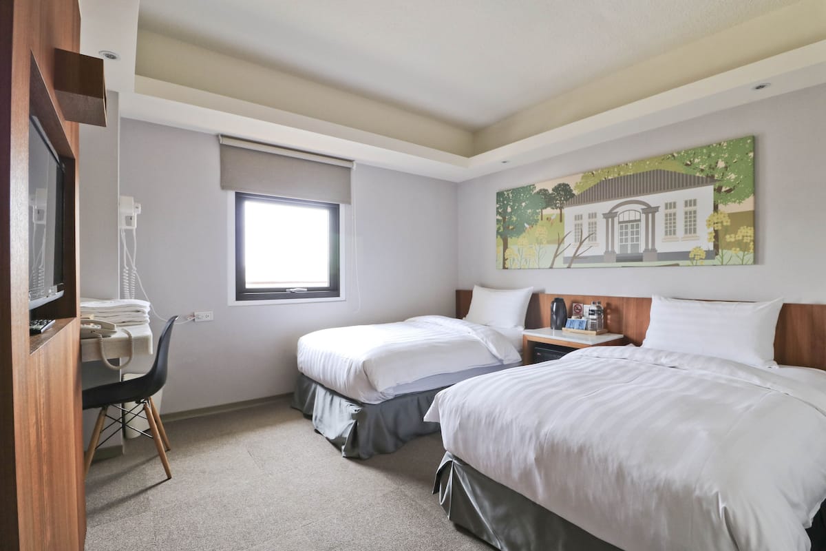 A hotel room with two beds, a desk with a chair, a window, and a painting on the wall.