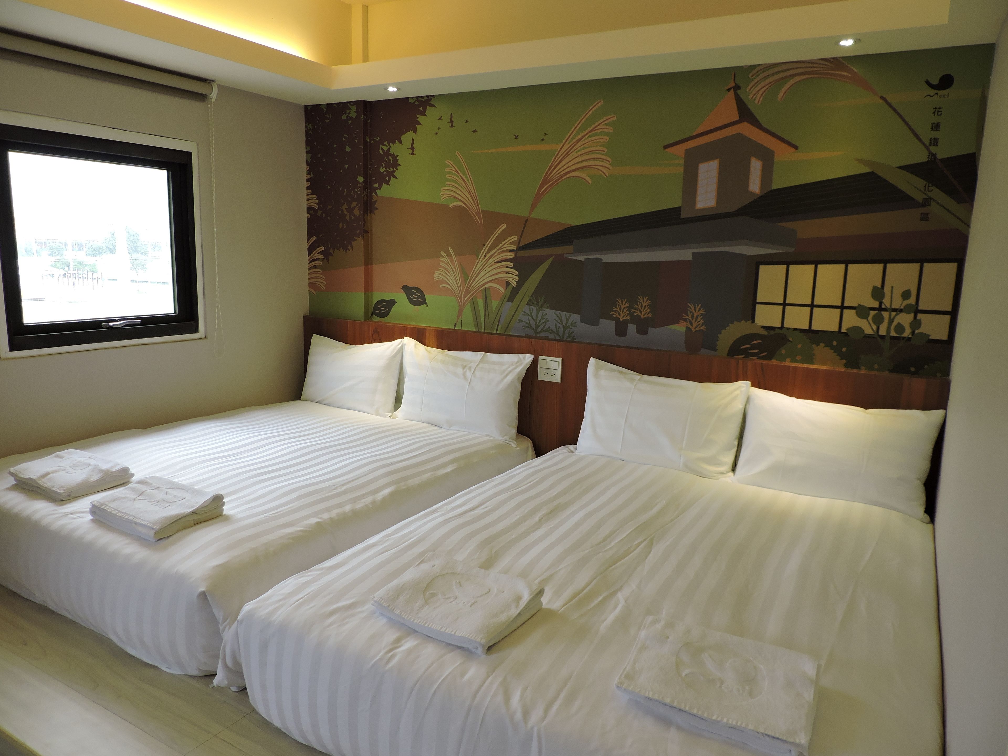 Habitación de hotel con dos camas, cada una con ropa de cama y toallas blancas, una ventana que ofrece vistas a zonas verdes y un mural en la pared que representa un paisaje con un edificio.