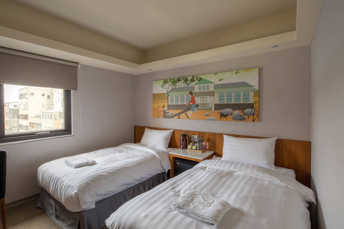 A hotel room with two beds, a painting on the wall, a window, and a small table with a kettle and other items.