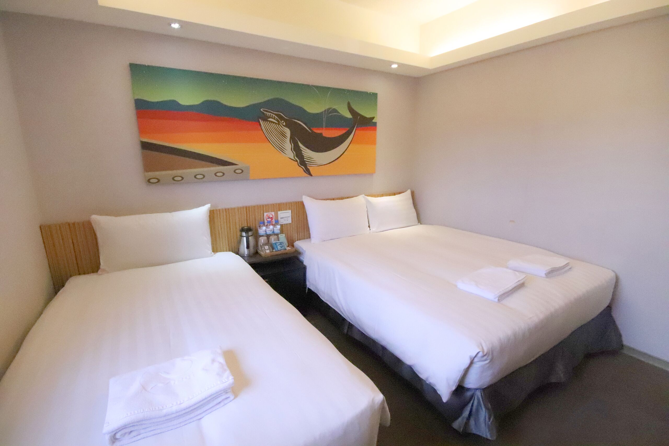Habitación de hotel con dos camas, un cuadro en la pared y una mesita de noche con artículos de aseo.