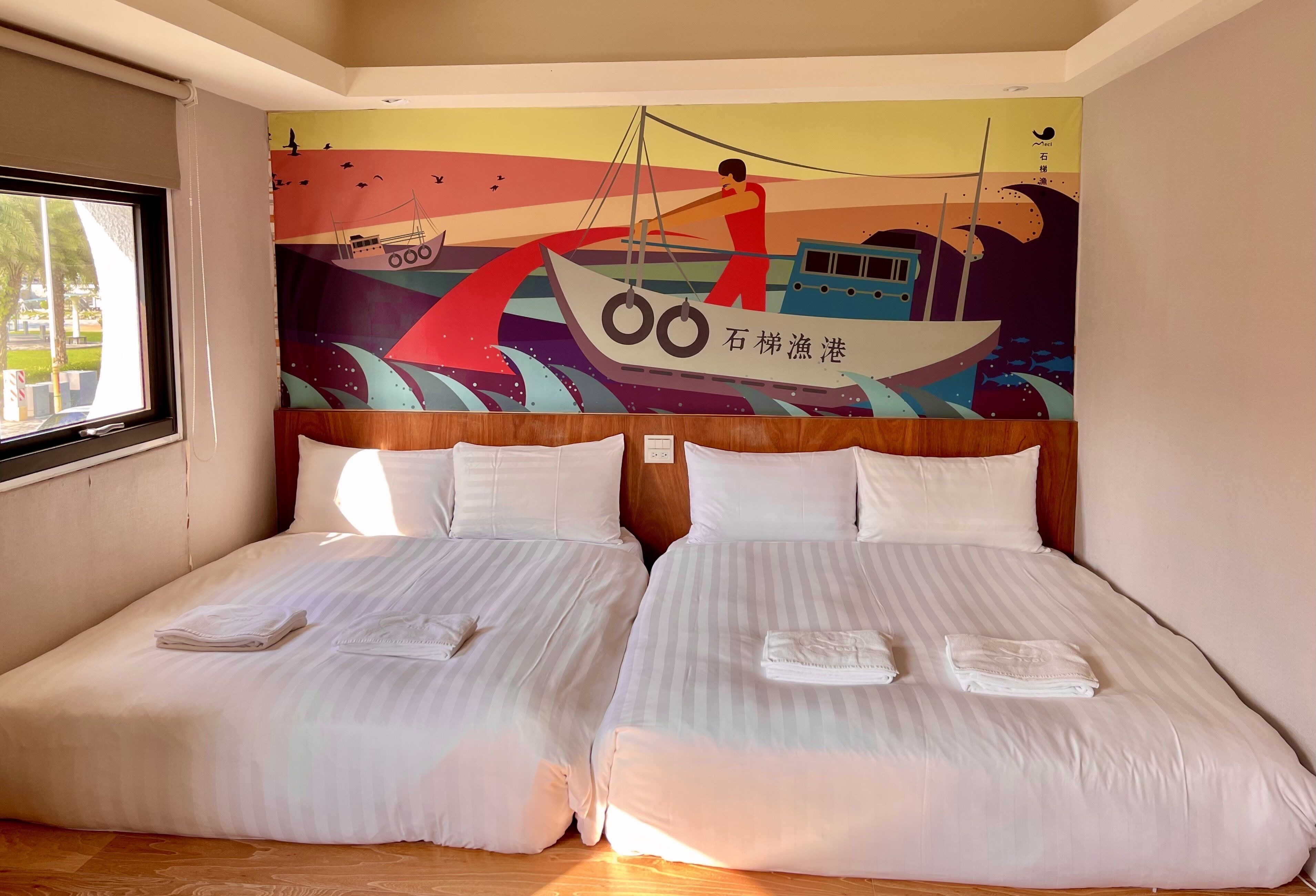 Habitación de hotel con dos camas, un mural de un barco en la pared y una ventana con vistas a árboles.