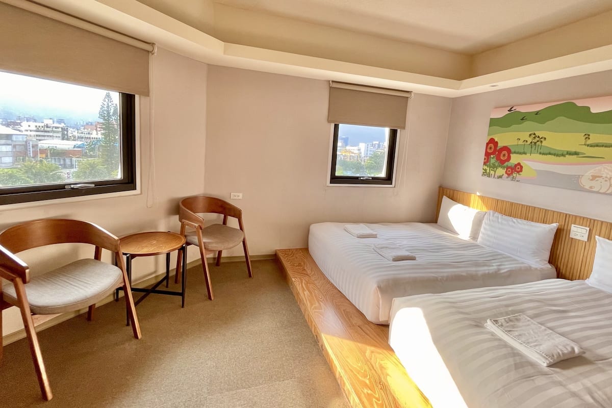 A hotel room with two beds, a small table, two chairs, and a view of the city through the windows.