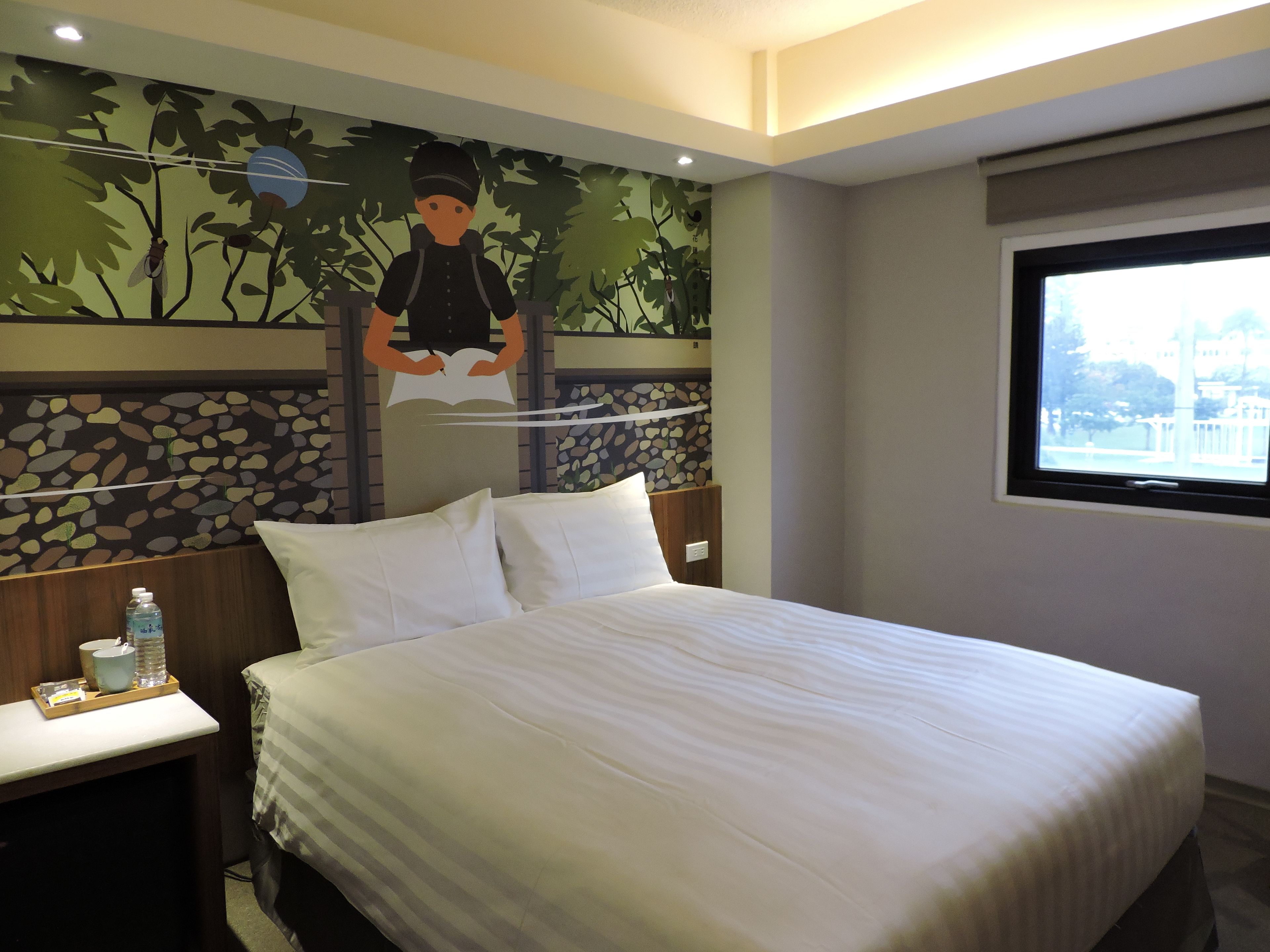 A hotel room with a bed, a nightstand with a water bottle and glasses, a window with a view, and a mural on the wall.