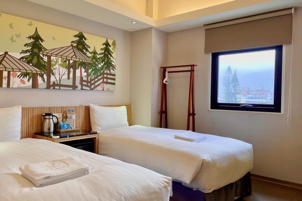 A hotel room with two beds, a window with a city view, a painting of trees, and a wooden nightstand with a kettle and bottles.