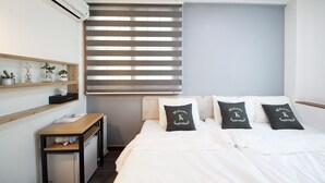 Ein Schlafzimmer mit einem Bett, zwei Kissen mit dem Aufdruck "Welcome K", einem Holztisch, einem kleinen Heizkörper und einem Fenster mit Jalousien.