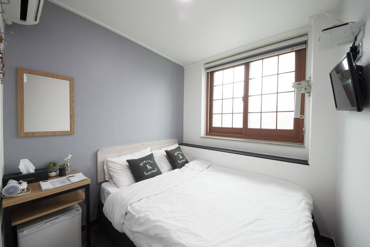 Een netjes ingerichte hotelkamer met een bed, een nachtkastje met lamp, een klein bureau met stoel, een raam met zonwering en een aan de muur bevestigde televisie.