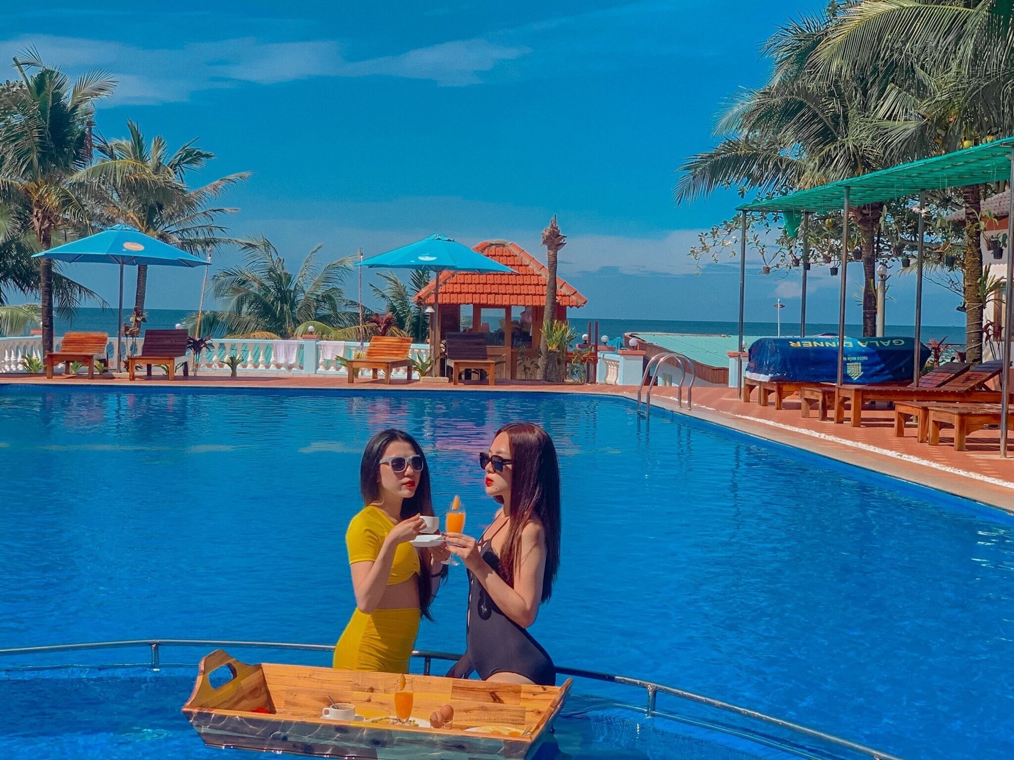 A pool area with lounge chairs and umbrellas, a small boat with a tray of food and drinks, and a view of the ocean.