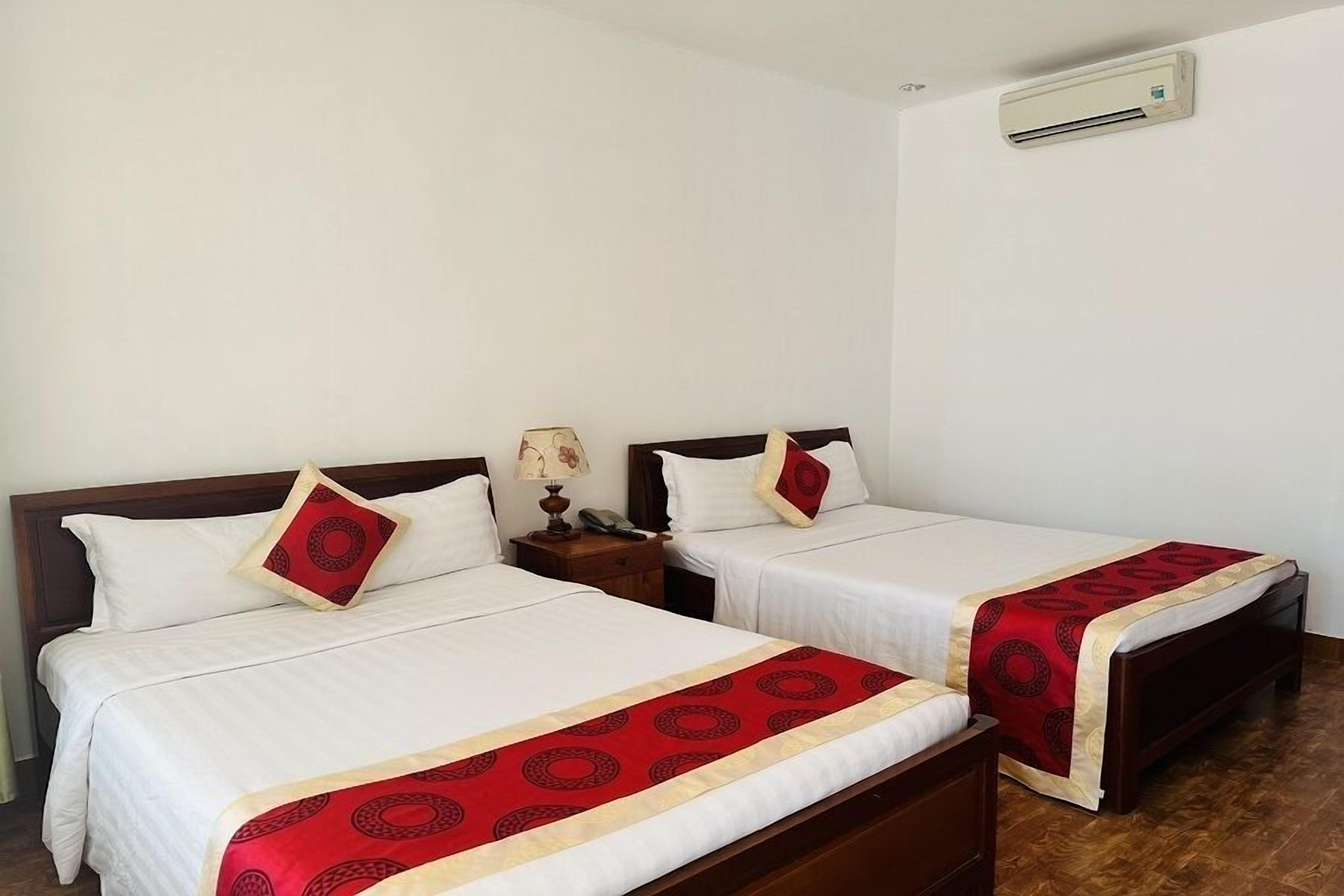 Two single beds with white linens and red decorative pillows, a wooden nightstand with a lamp, and an air conditioning unit on the wall.
