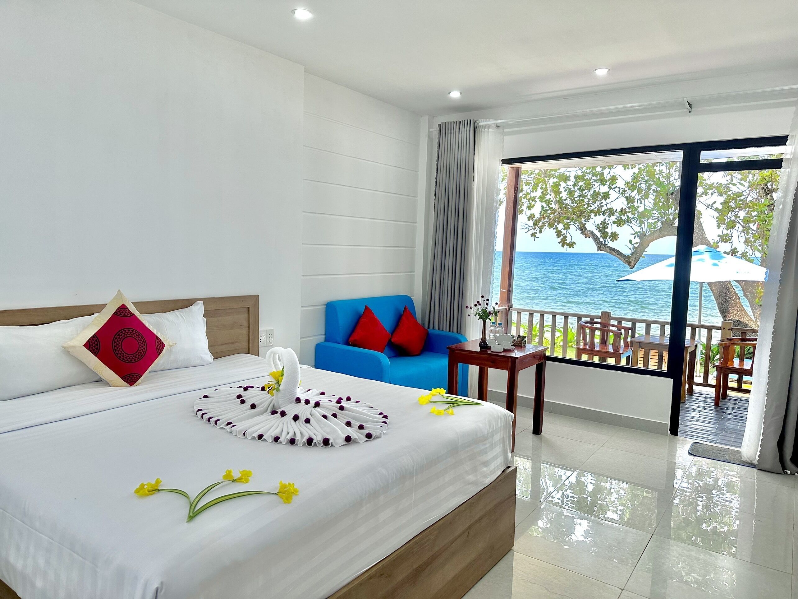 A hotel room with a bed, a blue sofa, a balcony with a view of the sea, and a table with a vase of flowers.