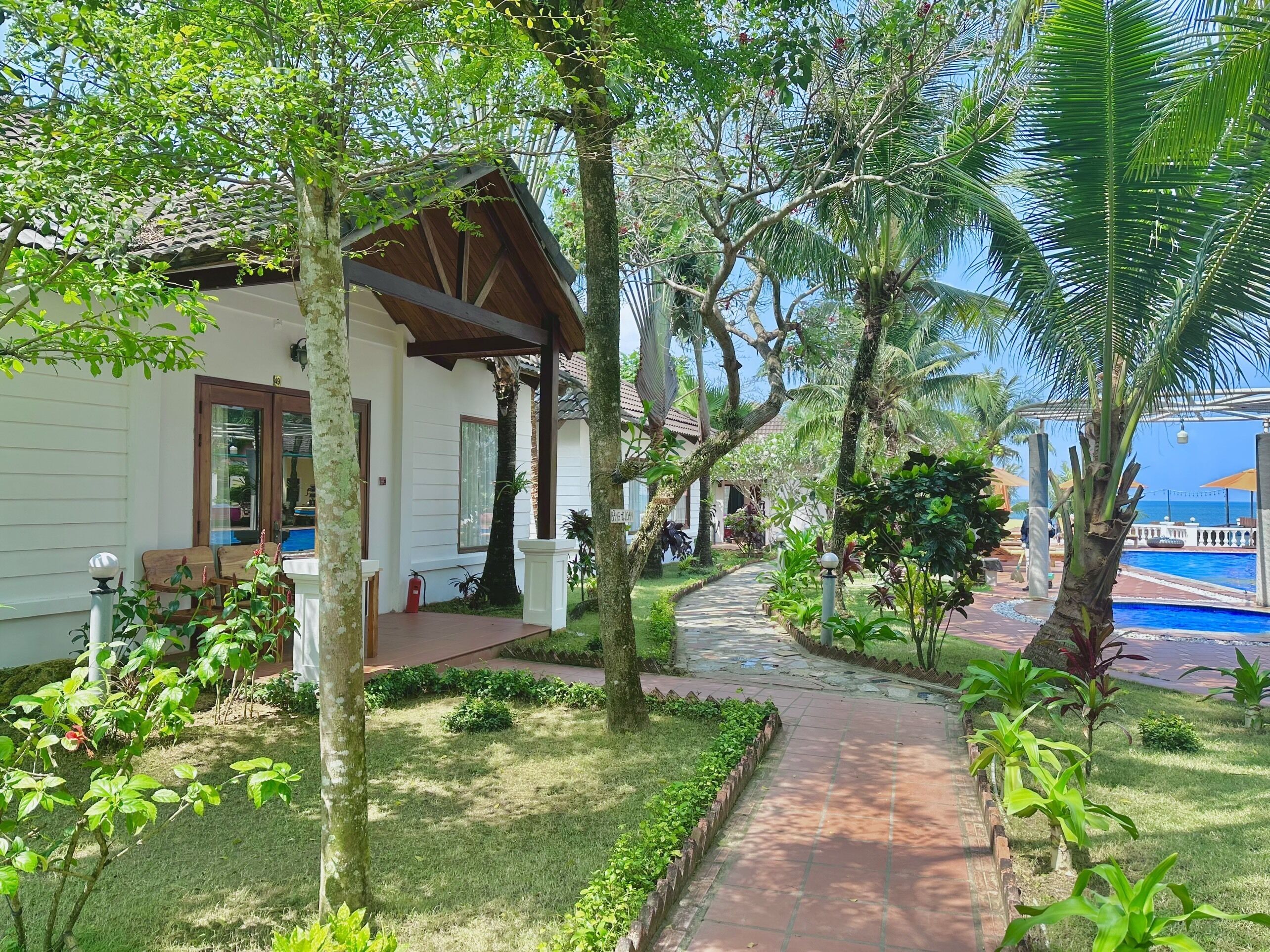 A pathway leading to a white building with a covered porch, surrounded by lush greenery and a pool area.