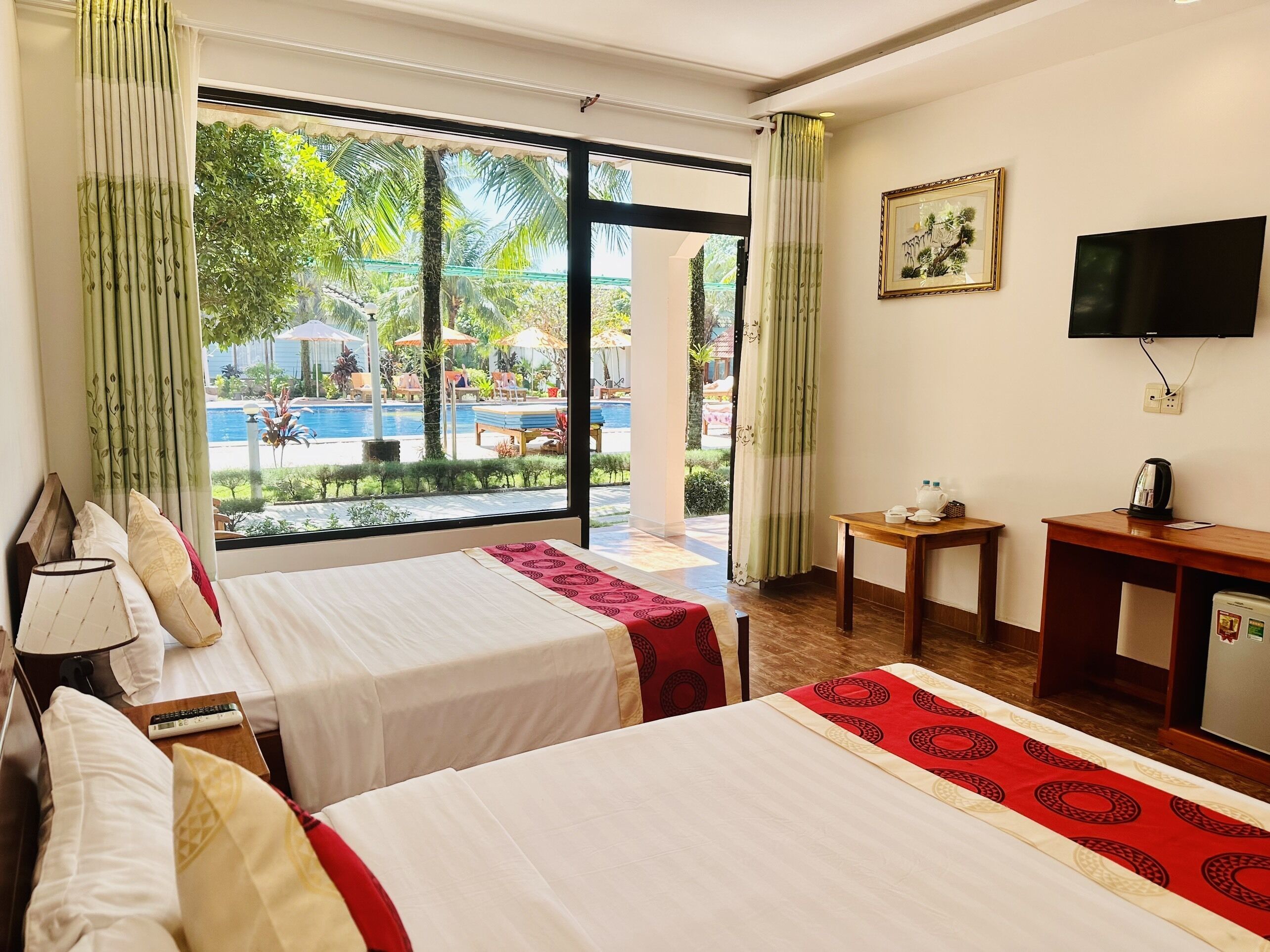 A hotel room with two beds, a TV, a small table with a kettle, and a view of a pool and greenery.