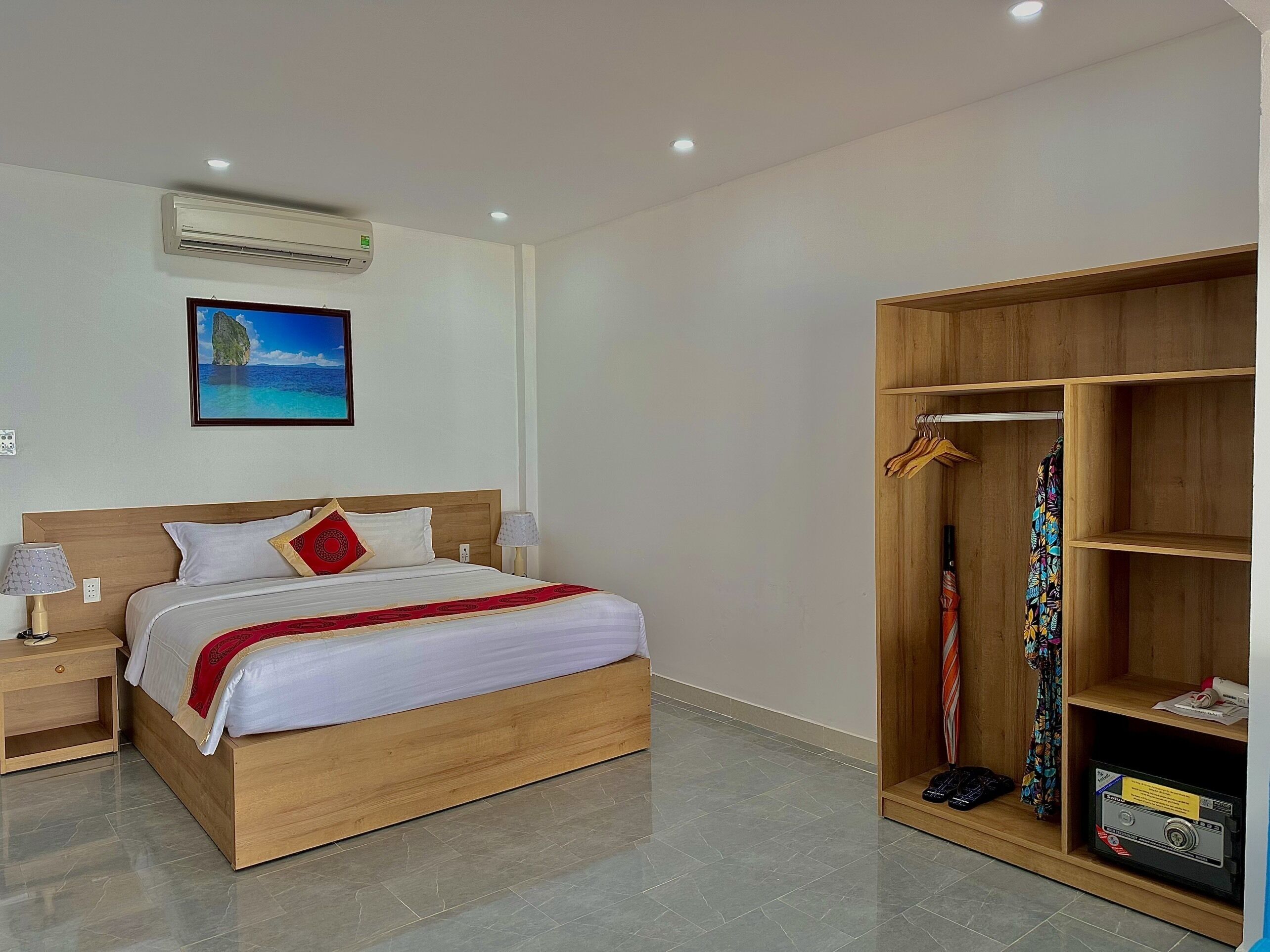 Hotelkamer met een houten bed, een kledingkast, een airconditioning, een nachtkastje, een lamp en een schilderij van een strand.