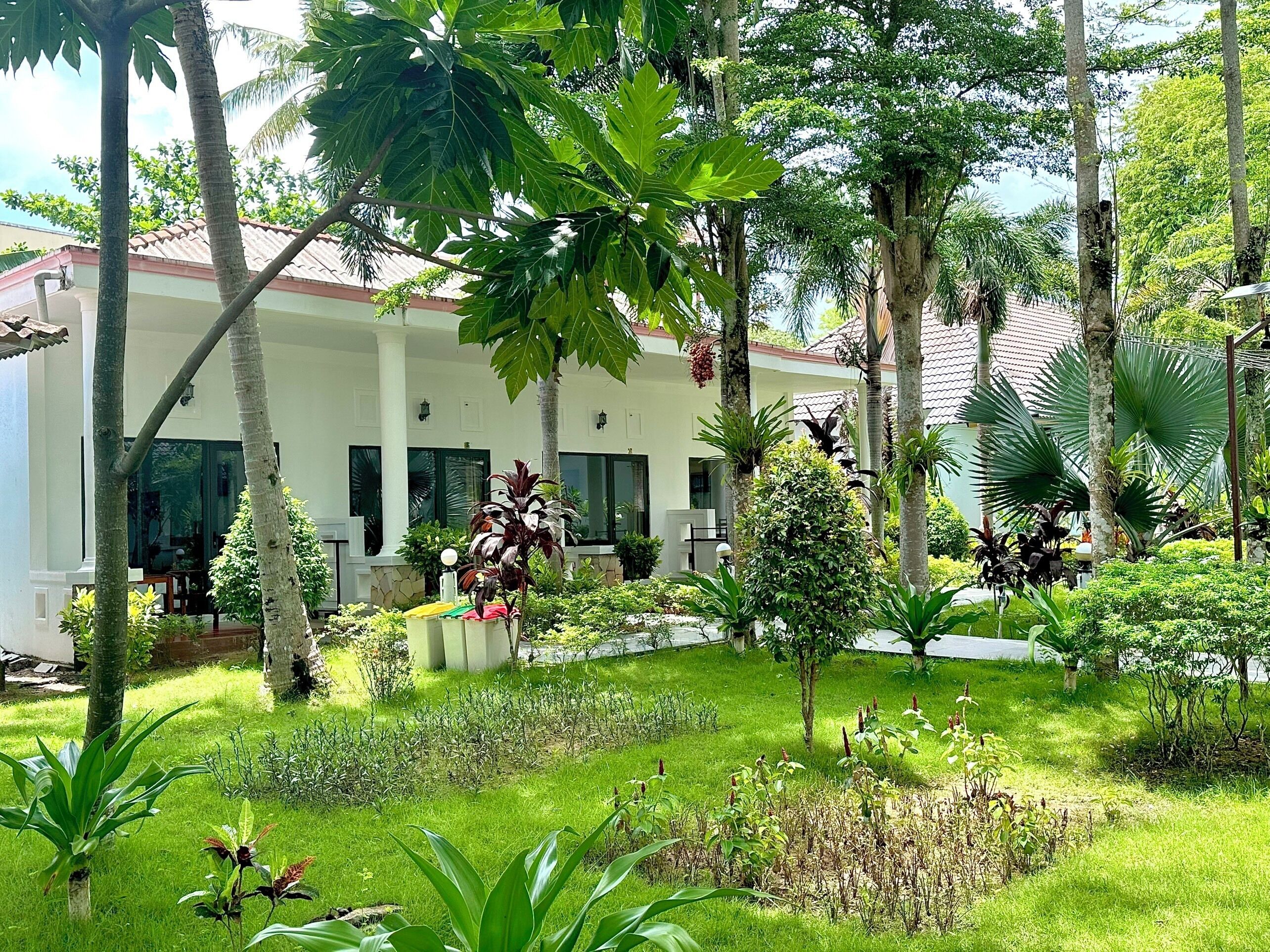 A lush garden with a variety of plants and trees, a white building with large windows, and a tiled roof.