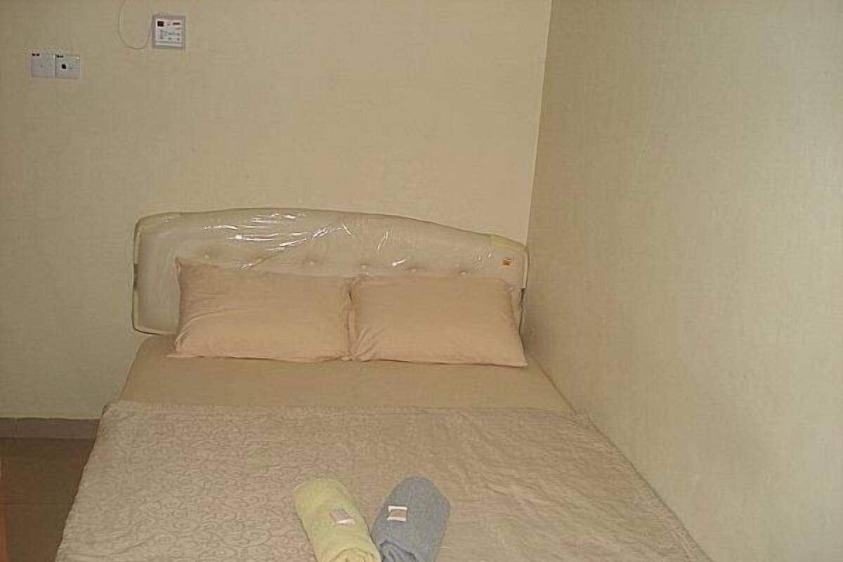 A small, single-bed room with a plain headboard, two pillows, and a bedspread. There are two rolled towels on the bed.