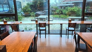 A restaurant with large windows offering a view of a garden and buildings. Tables and chairs are arranged for dining. - Cheonjiyeon Hotel (Seogwipo)