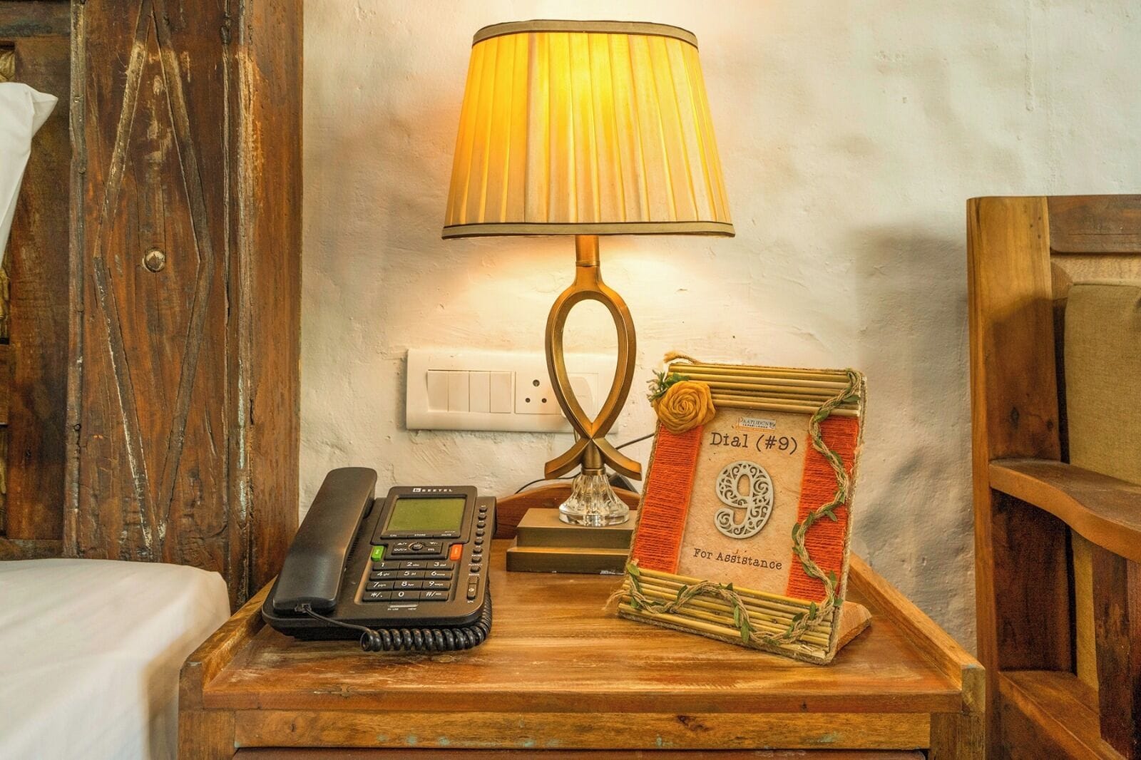 A wooden bedside table with a telephone, a lamp, and a framed card reading "Dial (#9) For Assistance".