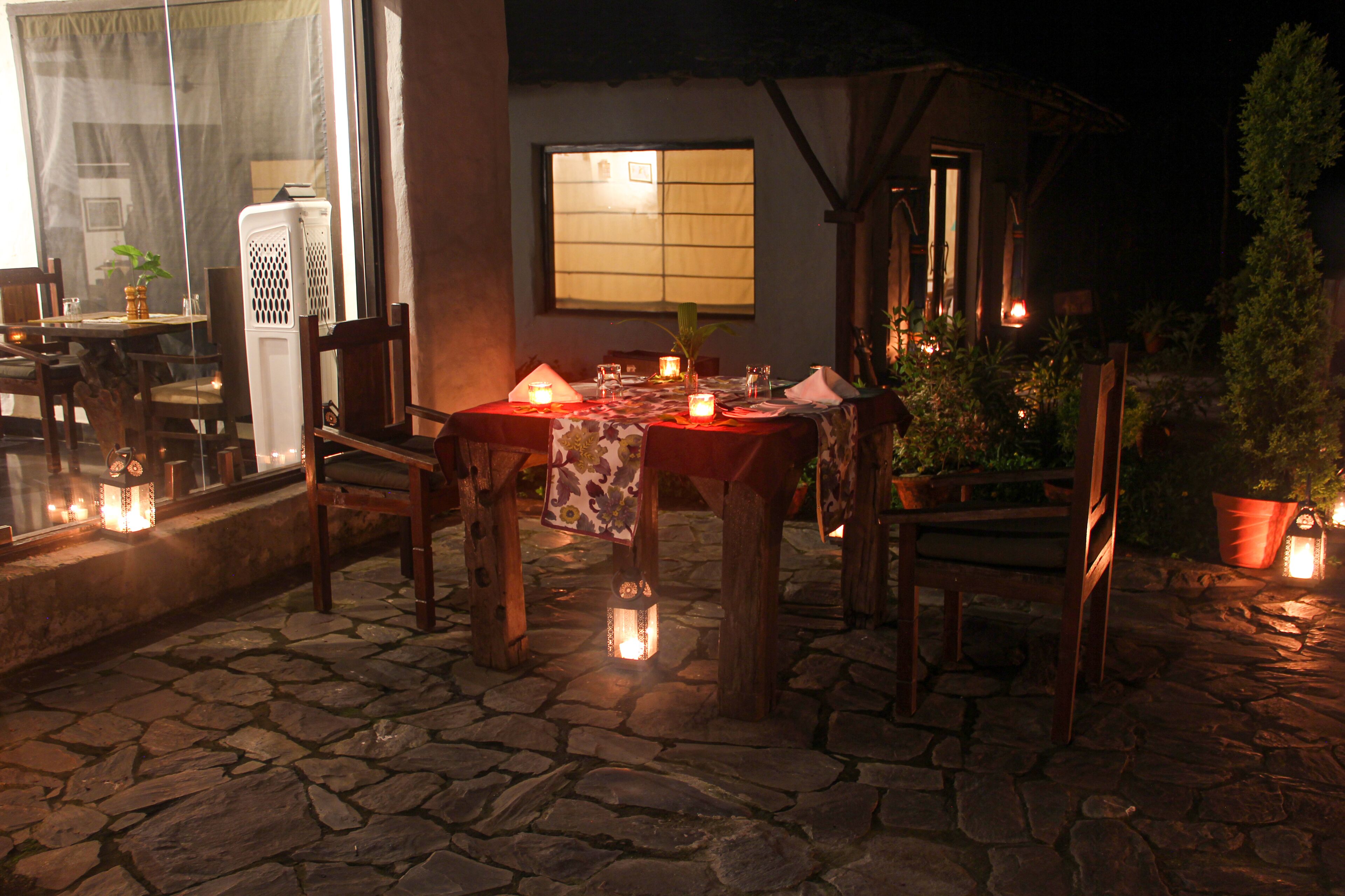 An outdoor dining area with stone flooring, wooden tables, and chairs, illuminated by lanterns.