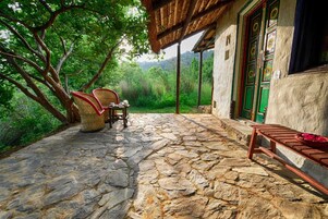 Un patio empedrado con muebles de mimbre y un banco de madera, rodeado de abundante vegetación y un edificio rústico con una puerta verde.