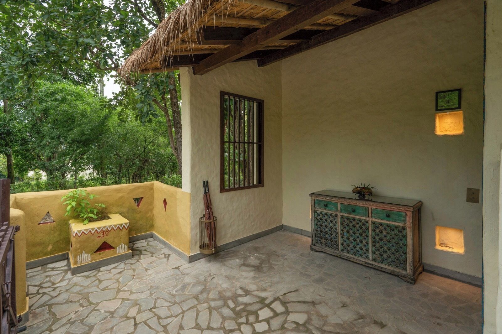 A room with a stone floor, a thatched roof, a window with bars, a small table with a plant, and a cabinet with decorative patterns.