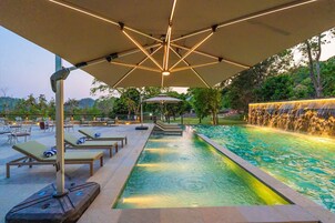 Una piscina con sillas de descanso y sombrillas, rodeada de vegetación y una cascada.