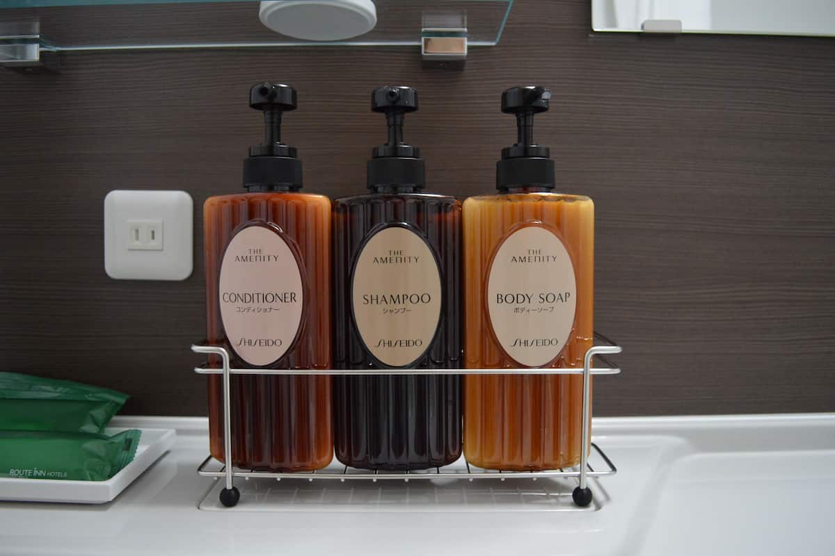 Un baño con un estante que contiene tres botellas de productos de tocador de la marca The Amenity: acondicionador, champú y jabón corporal.