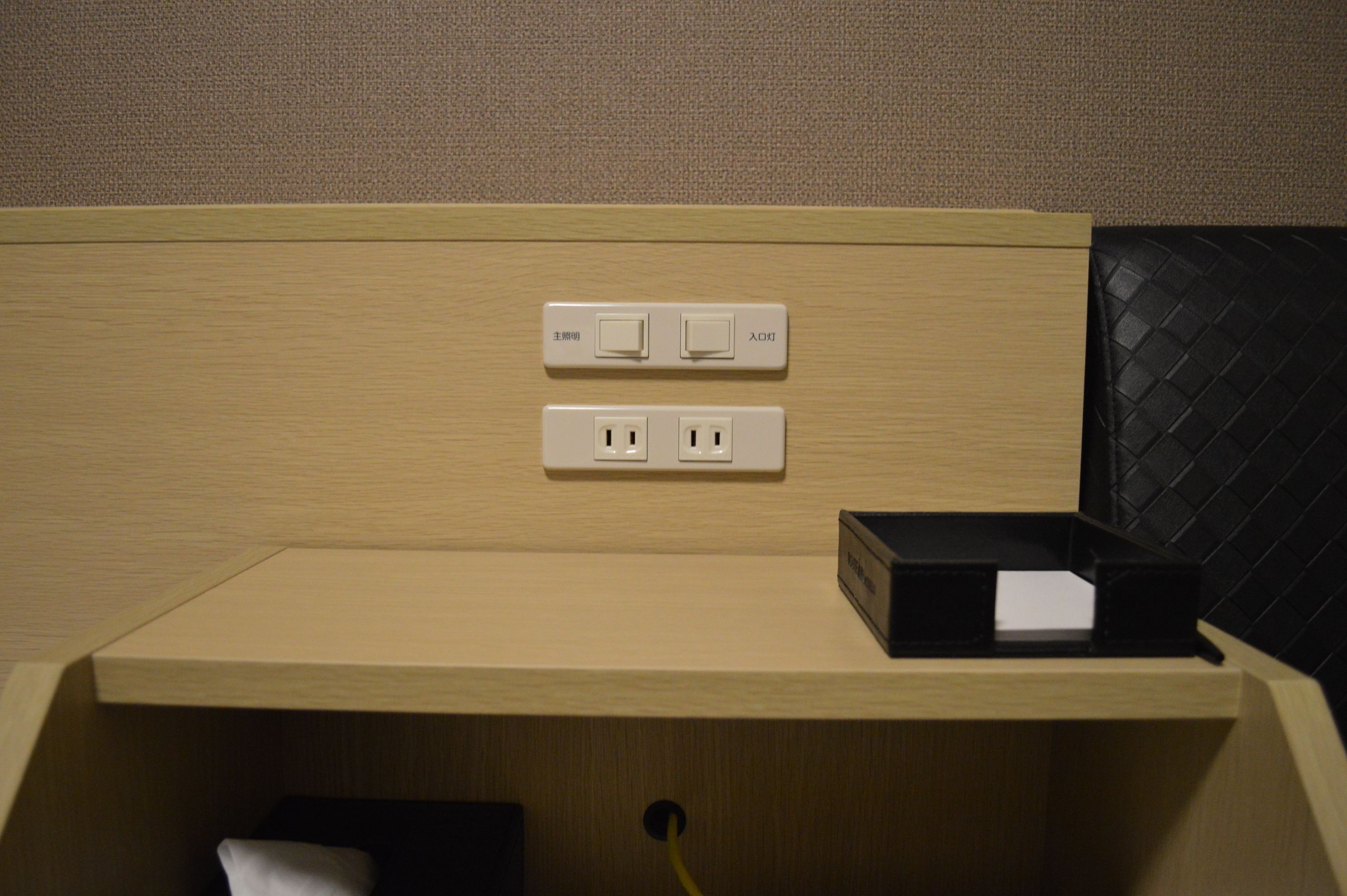 A hotel room with a wooden panel wall, a small wooden desk, and a black leather chair. There are two electrical outlets and a power strip on the wall.
