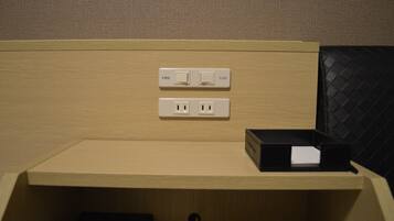 A hotel room with a wooden panel wall, a small wooden desk, and a black leather chair. There are two electrical outlets and a power strip on the wall.