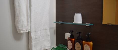 A bathroom with a white towel hanging on the wall, a glass shelf with a cup and three bottles of body soap.
