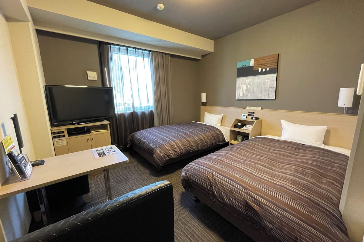 Habitación de hotel con dos camas, un escritorio, un televisor de pantalla plana, una mesita y una ventana con cortinas.