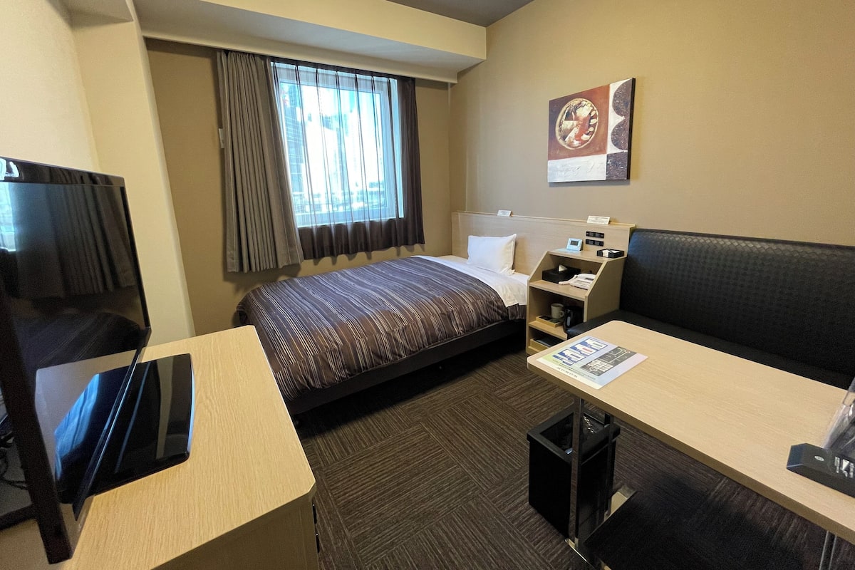 Habitación de hotel con cama, escritorio, televisor, ventana con cortinas, una obra de arte en la pared y un cubo de basura.