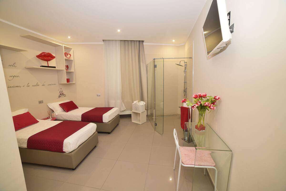 A hotel room with two beds, a glass shower enclosure, a small table with a chair, and a TV mounted on the wall.