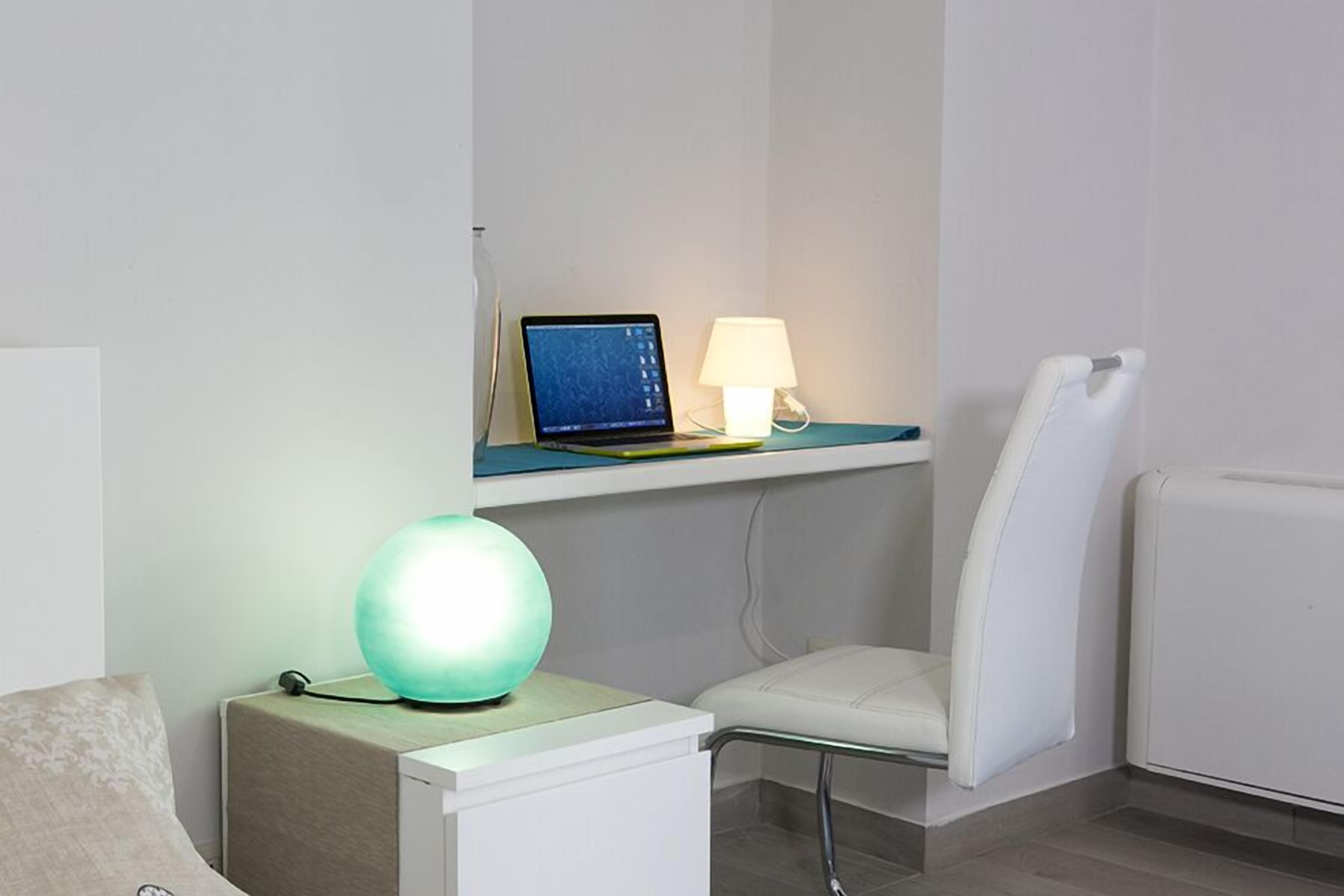 Un baño moderno con una computadora portátil sobre una repisa, una lámpara verde, una silla blanca y un radiador blanco.