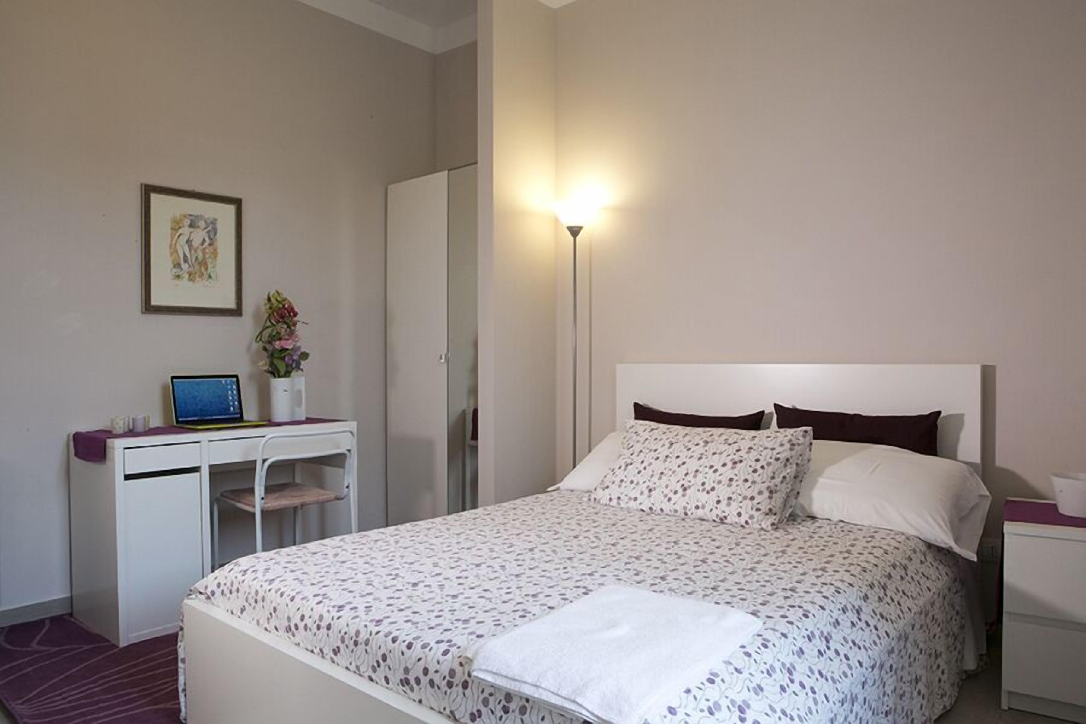 Un dormitorio con cama, escritorio con computadora portátil, mesita de noche, lámpara y un cuadro en la pared.