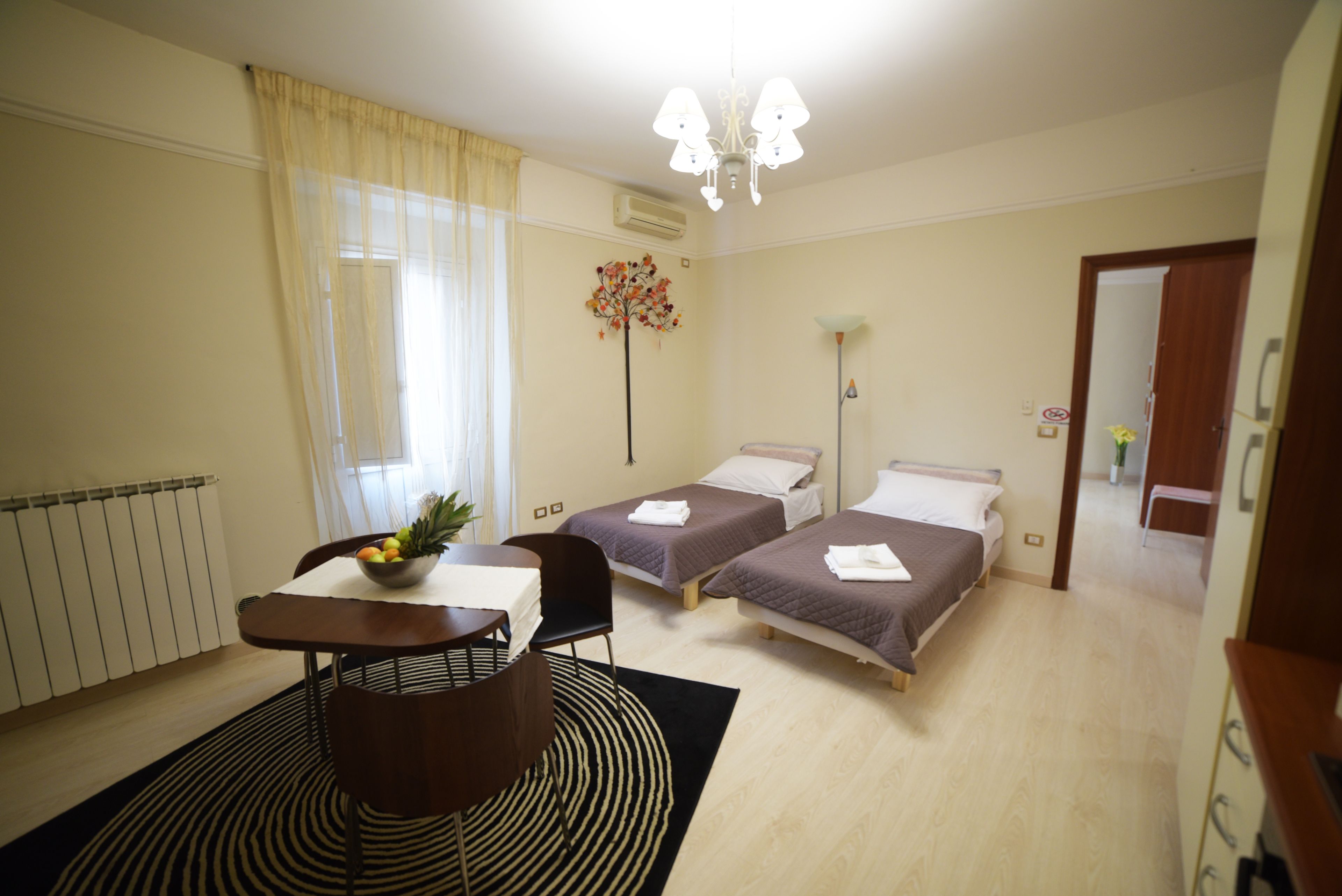 A hotel room with two beds, a dining table with chairs, a black and white striped rug, a flower vase, and a chandelier.