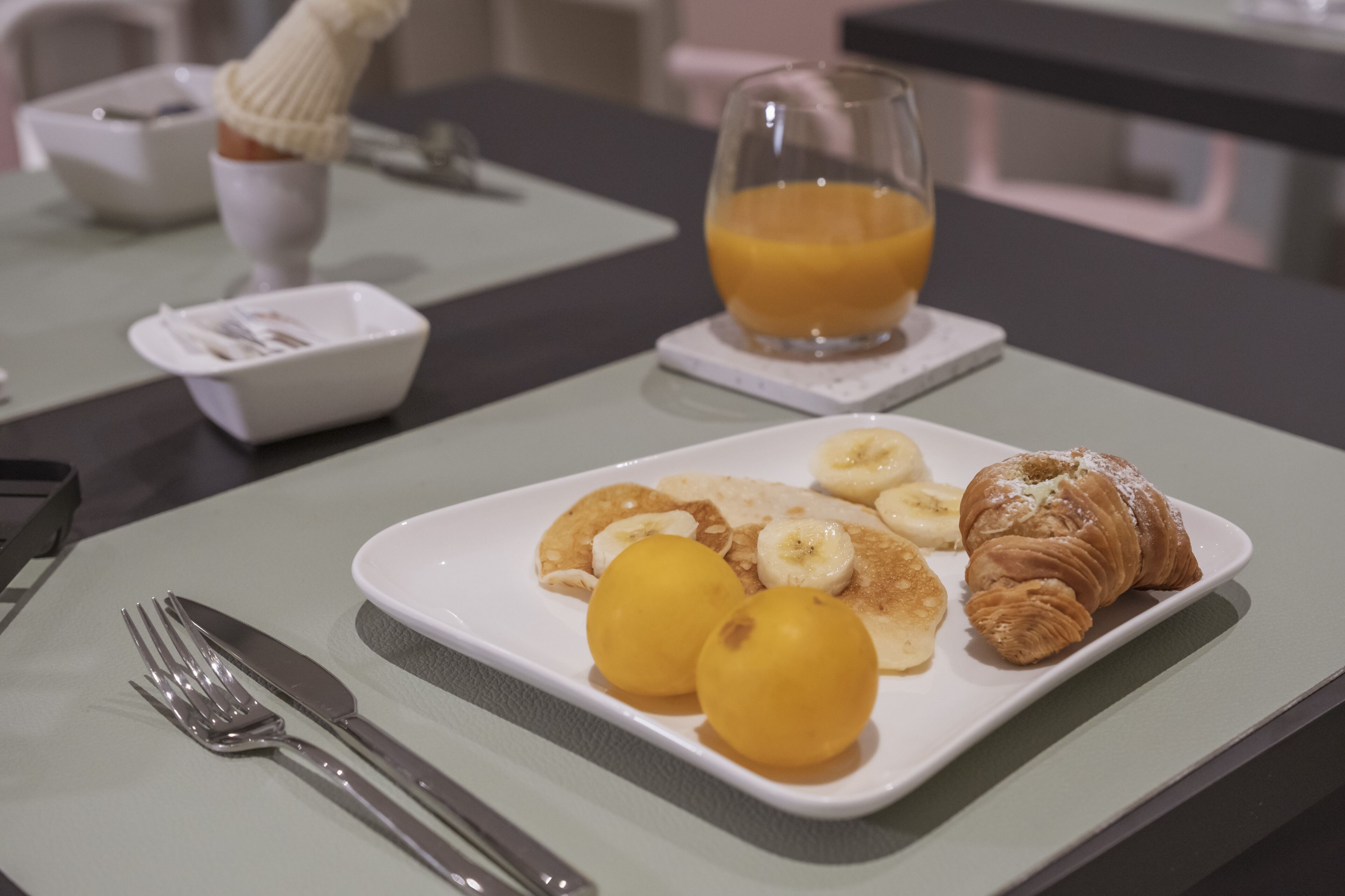 Une table dressée pour le petit-déjeuner, avec une assiette de nourriture comprenant des bananes, des croissants et un verre de jus d’orange.