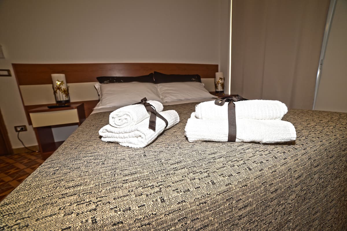 A neatly made bed with white linens and brown ties, a wooden headboard, and a nightstand with a lamp and a vase with flowers.