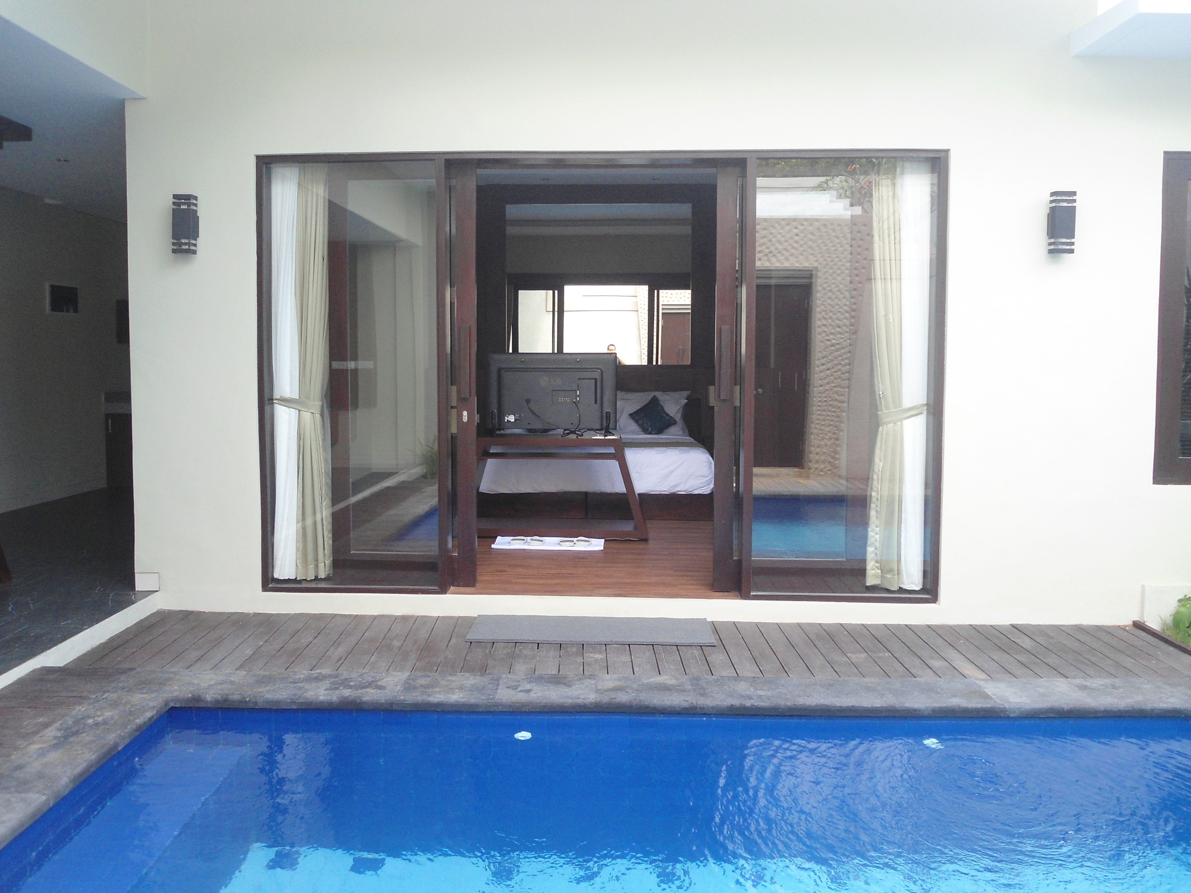 A modern hotel room with a large sliding glass door leading to a balcony, a bed with a view, and a pool area.