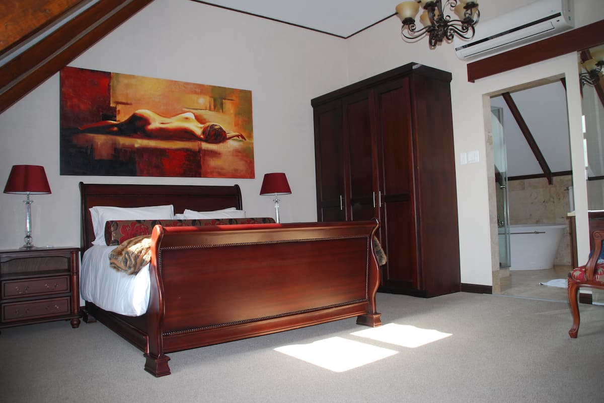 A bedroom with a wooden bed, a large painting, a wardrobe, and a bathroom visible through an open door.