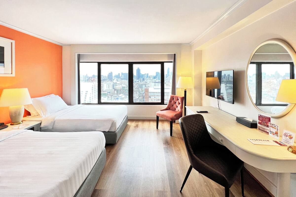 Une chambre d’hôtel avec deux lits, un bureau, une chaise, une télévision et une grande fenêtre donnant sur la ville.