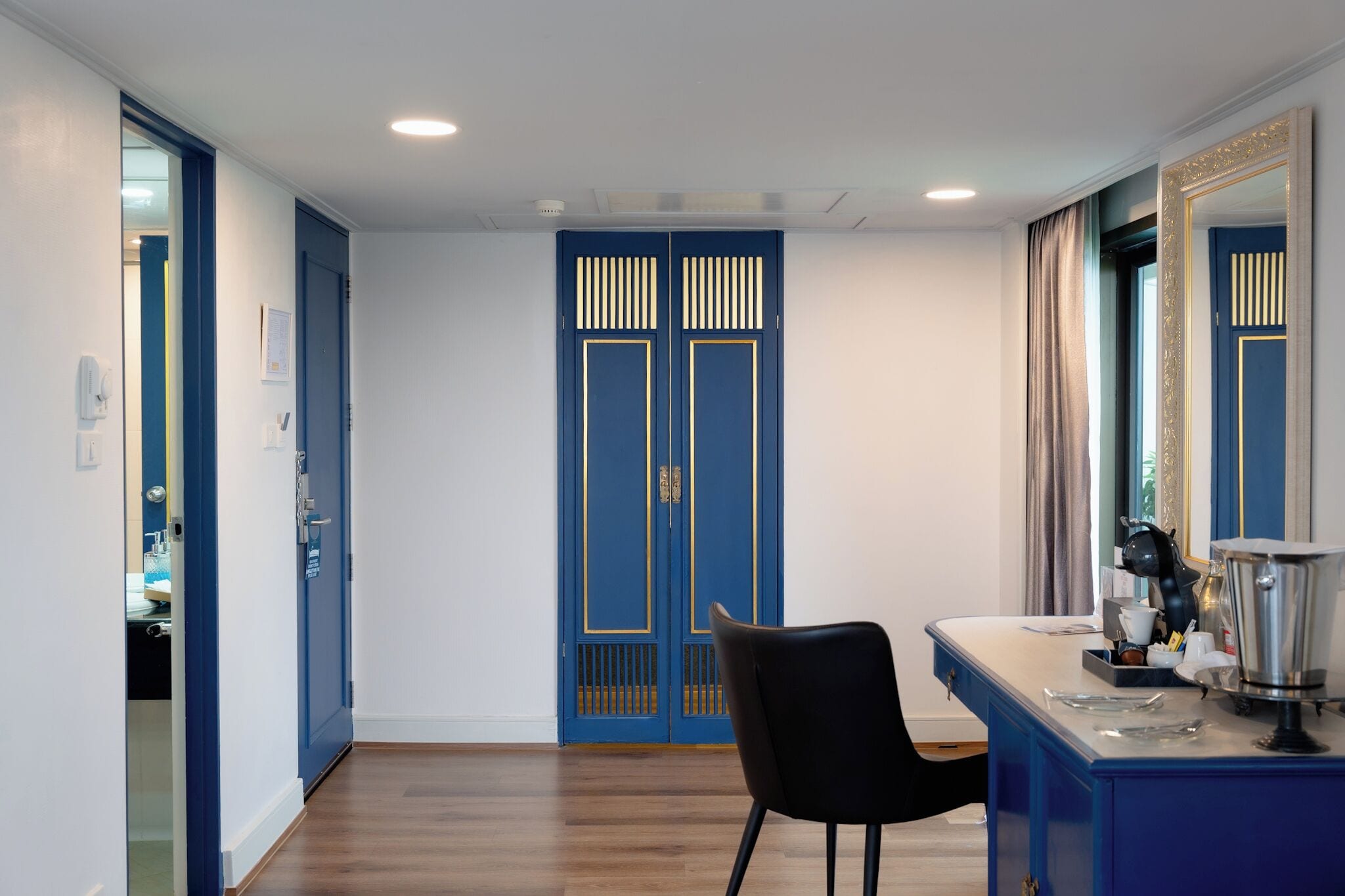A room with a blue door, a black chair, a desk with a coffee maker, and a mirror.