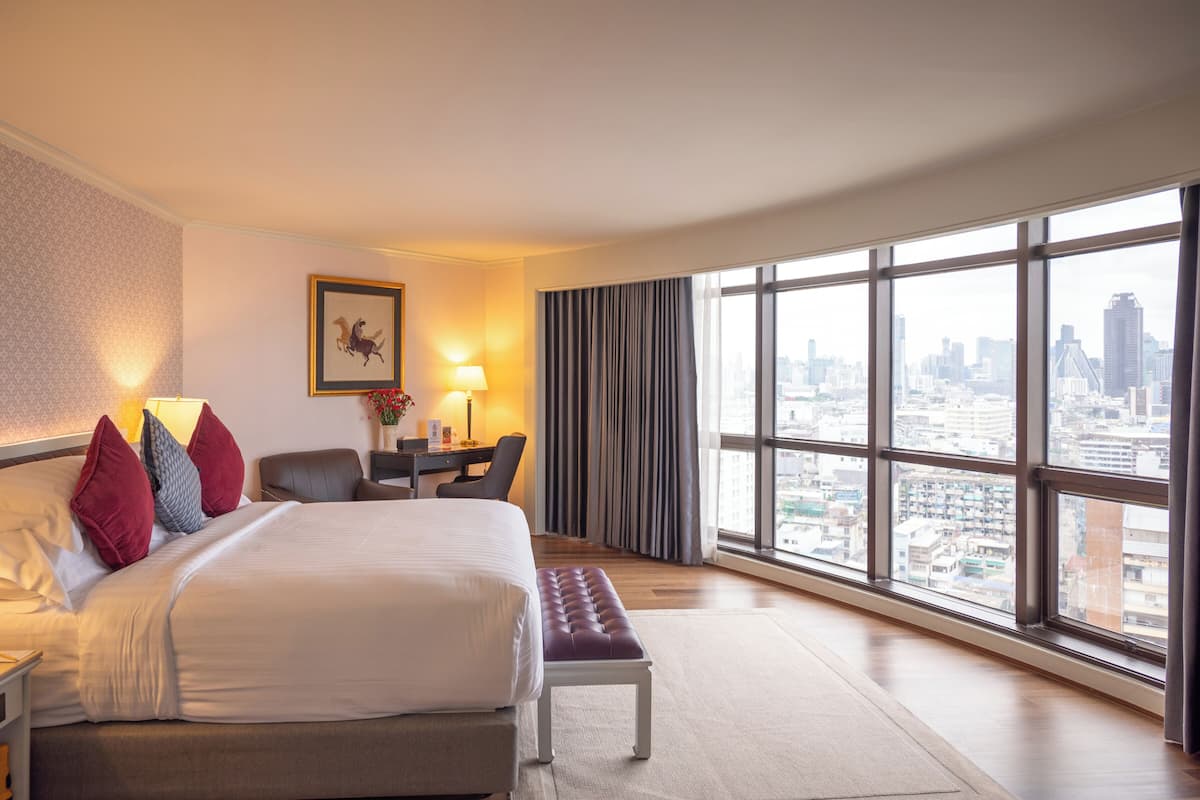 Une chambre d’hôtel dotée d’un grand lit, d’un bureau, d’une chaise, d’un banc et offrant une vue sur le paysage urbain grâce à de grandes fenêtres.