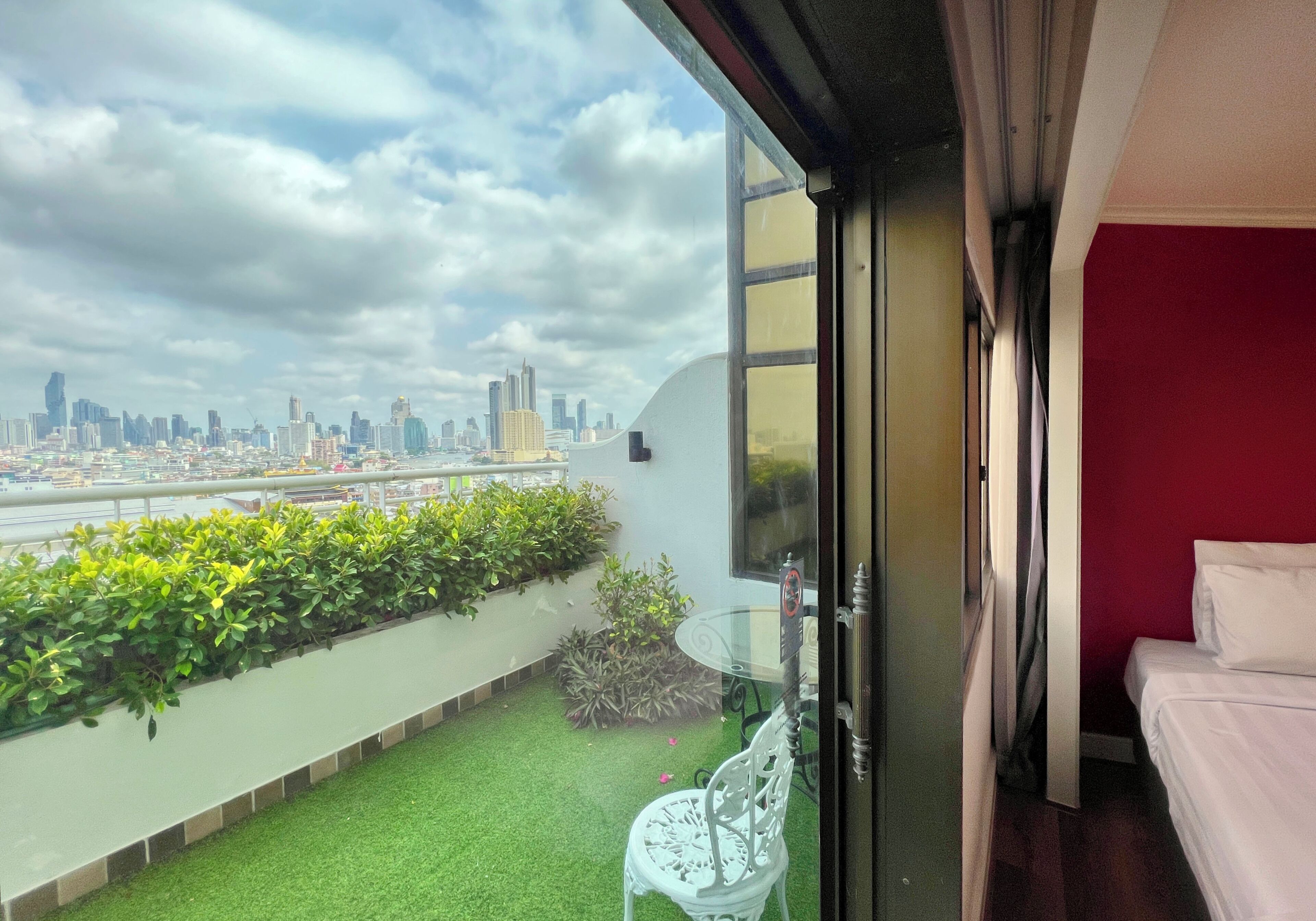 A balcony with a city view, a small table with a chair, and a bed with a red wall.