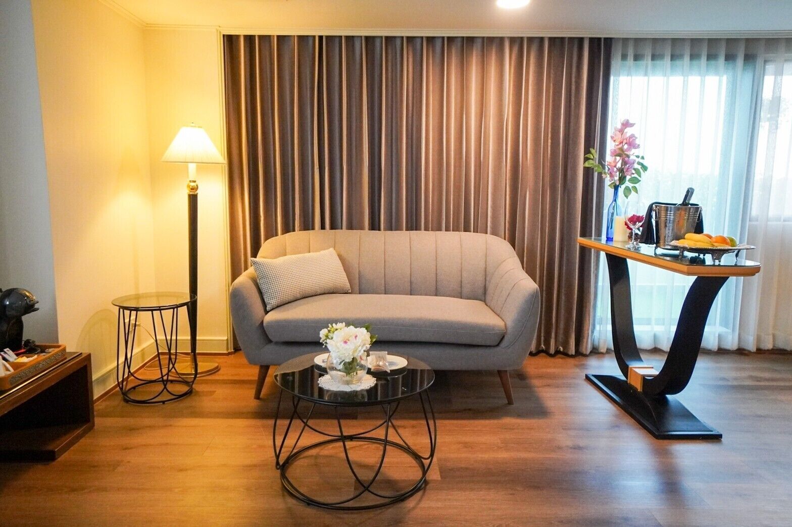 A living room with a sofa, a round coffee table with flowers, a lamp, and a dining table with fruits.