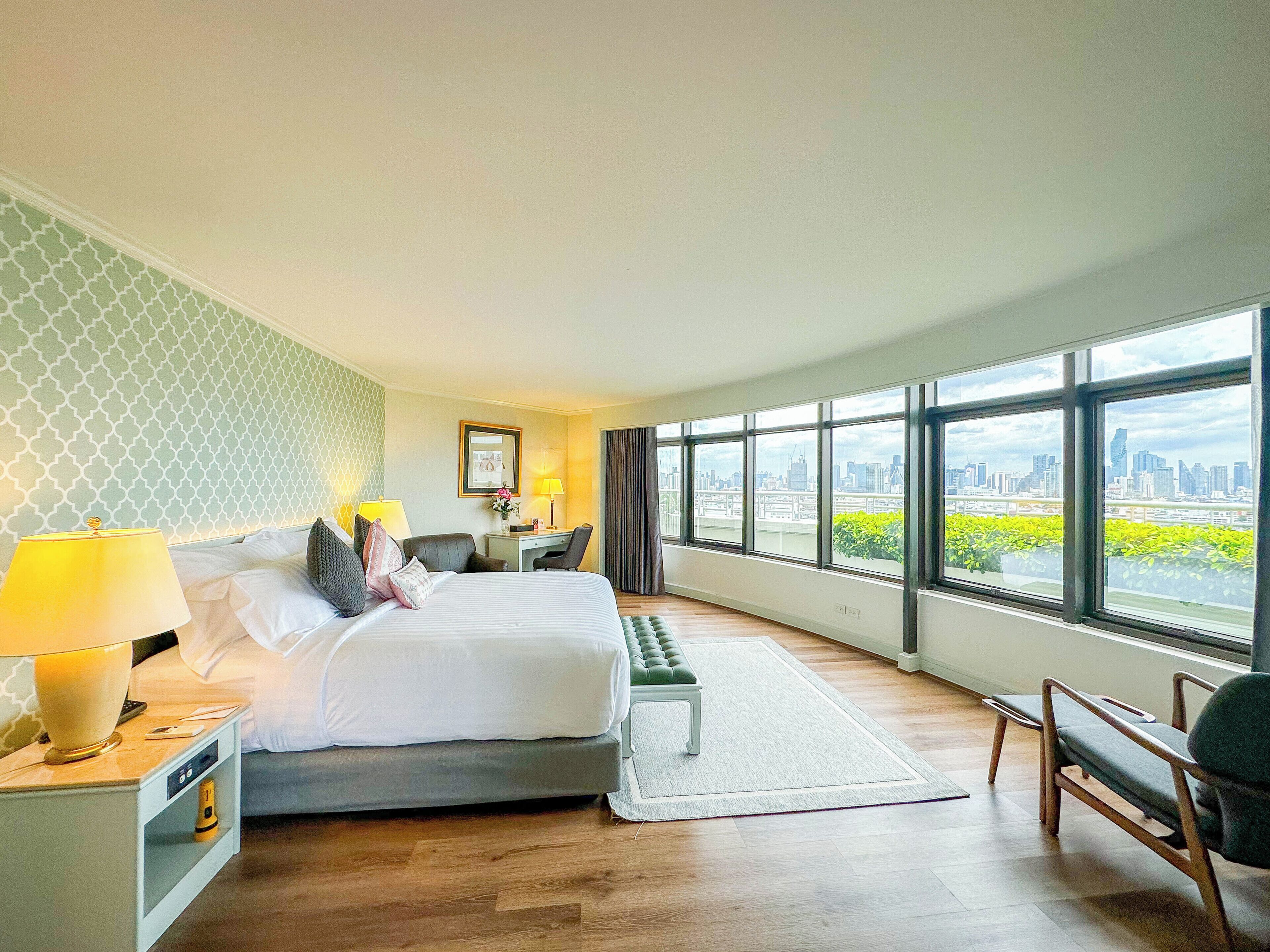 A hotel room with a bed, bedside lamps, a desk, a chair, and large windows offering a city view.