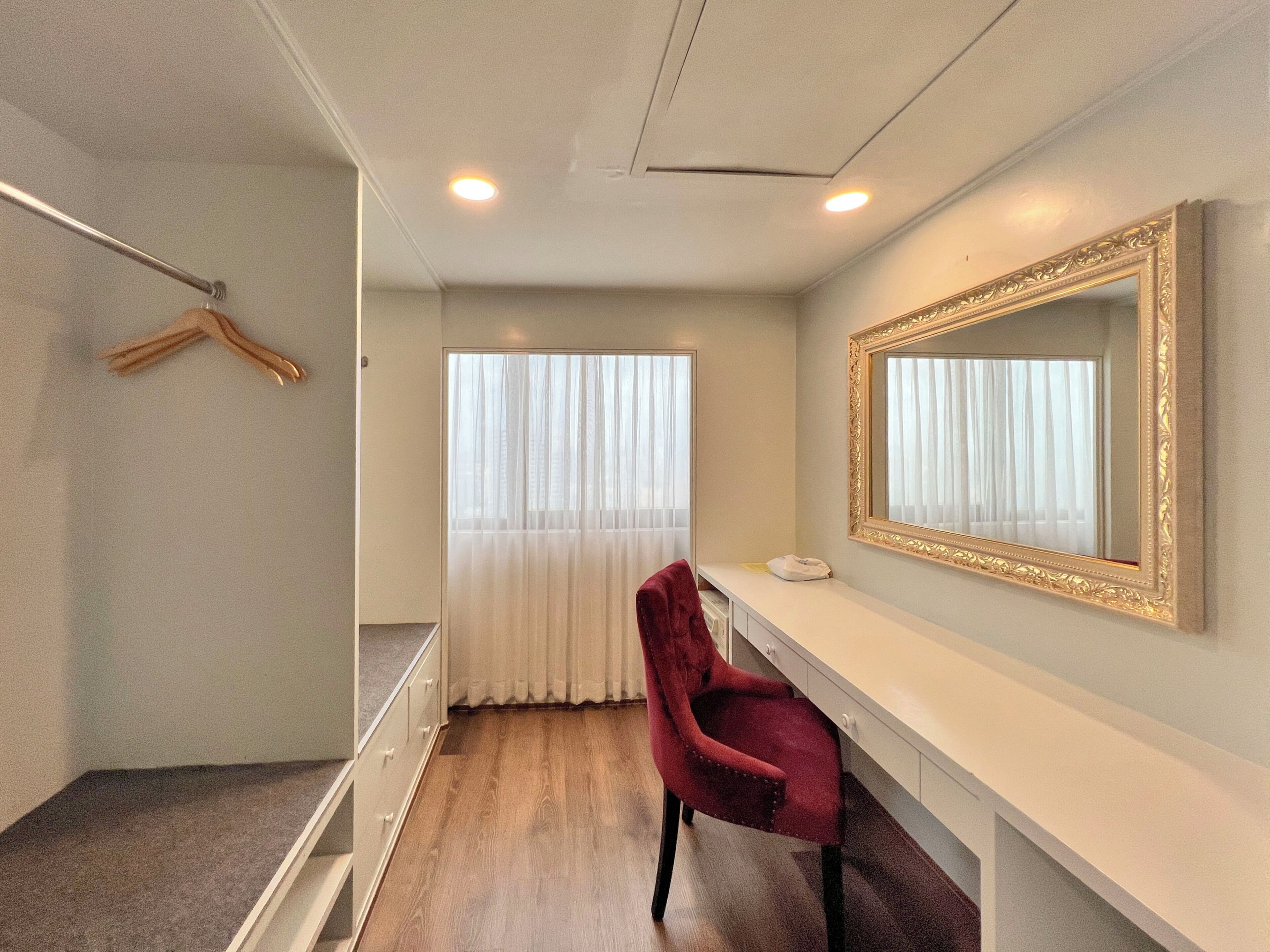 A hotel room with a vanity, a mirror, a red chair, a coat rack, and a window with curtains.