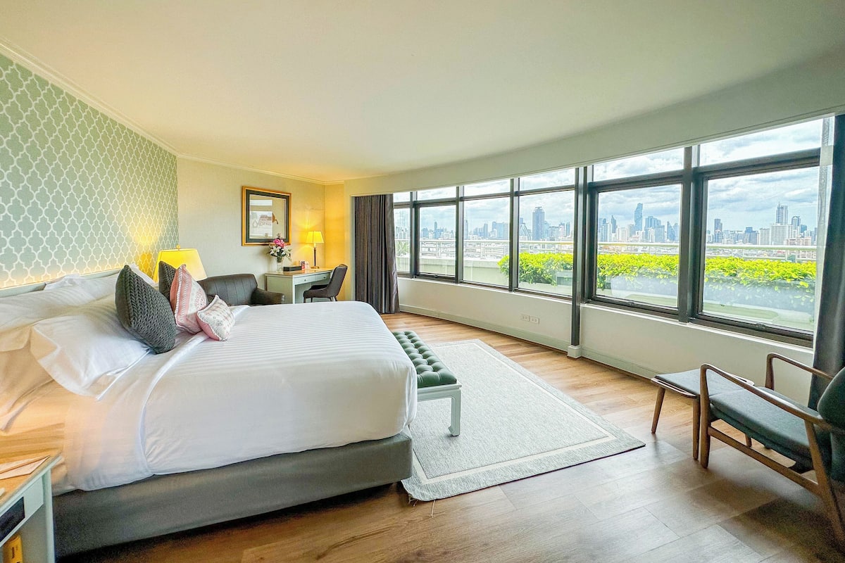 Une chambre d’hôtel équipée d’un lit, d’un bureau, d’une chaise et de grandes fenêtres donnant sur la ville.