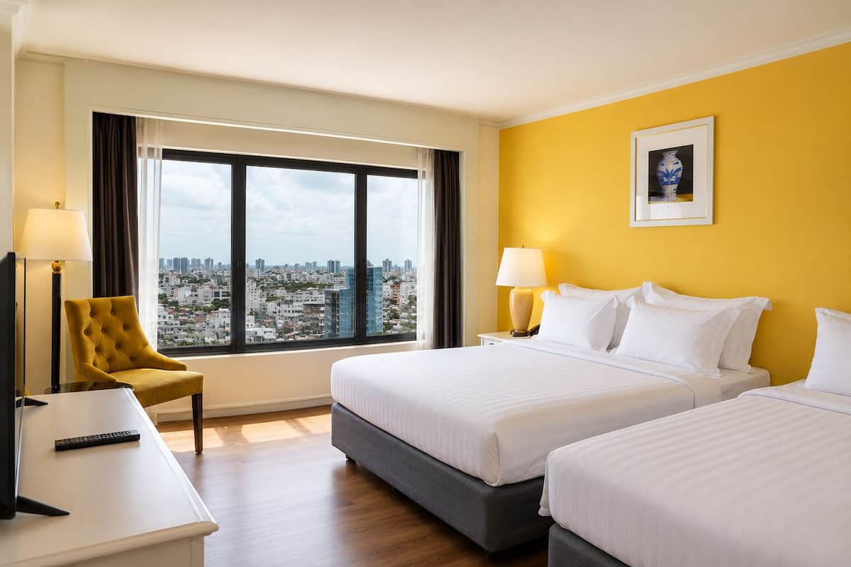 Une chambre d’hôtel avec deux lits, une chaise, un bureau, une lampe et une grande fenêtre donnant sur la ville.