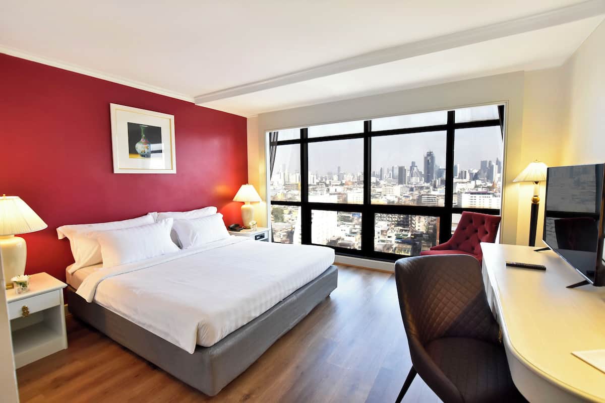 Une chambre d’hôtel comprenant un lit, un bureau, une chaise, une télévision, une fenêtre donnant sur la ville et un tableau encadré au mur.